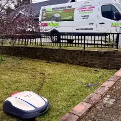 En robotgräsklippare från Honda står på en gräsmatta. En vit servicebil med texten "TrädgårdSmart" och "gardena blinkar blått" står parkerad bakom ett staket.