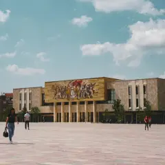 Plac w centrum Tirany w Albanii, z monumentalnym budynkiem ozdobionym mozaiką i ludźmi spacerującymi po rozległym, wybrukowanym placu.