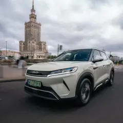 Biały SUV z logo chińskich samochodów BYD jedzie ulicą w Warszawie, z Pałacem Kultury w tle.