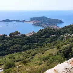 Widok z góry na wybrzeże Morza Śródziemnego, z licznymi jachtami na wodzie i zielonymi wzgórzami porośniętymi drzewami. W oddali widać półwysep Cap Martin.