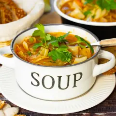 Herzhafte Kohlsuppe, low carb, in einem weißen Topf mit der Aufschrift "SOUP", garniert mit Petersilie. Dazu gibt es Vollkornbrot und Karotten.