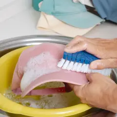 Ręce szorują różową czapkę z daszkiem niebieską szczotką w żółtej misce z wodą i pianą. Dowiedz się, jak wyczyścić czapkę z daszkiem.