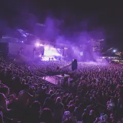 Tłum ludzi na koncercie pod fioletowym niebem, światła sceny i dym tworzą **motto innego świata**.