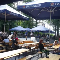 Ein sonniger Tag im Paulaner Biergarten. Gäste sitzen an Holztischen unter großen Sonnenschirmen und genießen die Atmosphäre.