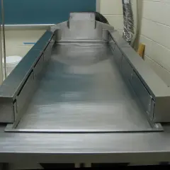 Stół do sekcji zwłok ze stali nierdzewnej, gotowy do pracy. Obok wisi tablica z anatomią człowieka.