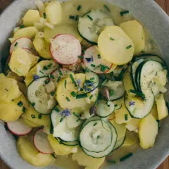 Kartoffelsalat mit Gurken, Radieschen und Schnittlauch, garniert mit kleinen blauen Blüten. Ein frischer Genuss auf Holztisch.
