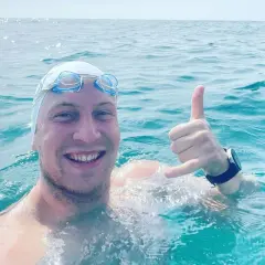 Uśmiechnięty pływak w czepku i goglach, z ręką w geście "hang loose", w trakcie przepłynięcia Bałtyku wpław.