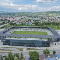 Nowoczesny stadion Sandecji z zieloną murawą i trybunami, otoczony miastem i wzgórzami.