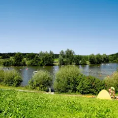 Sonniger Tag am Aichstruter Stausee. Menschen planschen im Wasser, baden und entspannen sich am Ufer unter bunten Sonnenschirmen.