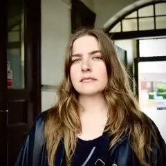 Miriam Ingrid, med sitt långa, vågiga hår och gröna ögon, blickar ut från en mörk port.