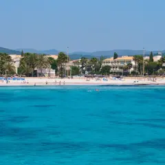 Türkisblaues Meer vor Sa Coma Altstadt mit Palmen und Hotels. Menschen genießen den Strand und das Wasser.