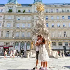 Due bambini si abbracciano davanti alla Colonna della Peste a Vienna. Altri bambini passeggiano sullo sfondo.