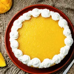 Sernik mango na zimno, ozdobiony bitą śmietaną, obok kawałka mango i łopatki do ciasta.