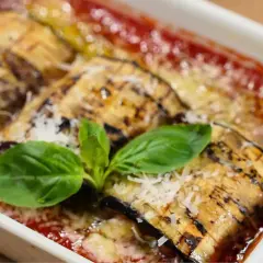 Rollo de pastel de berenjenas a la parmesana, gratinado con queso y bañado en salsa de tomate, decorado con hojas de albahaca fresca.