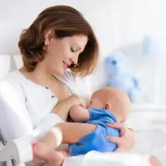 Uśmiechnięta mama karmi piersią noworodka w białym fotelu. Delikatne chwile bliskości, choć czasem pojawia się ulewanie u noworodka.