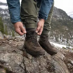 A hiker laces up sturdy brown boots on a rocky mountain trail. The question of should you size up in boots is crucial for comfort on such terrain.