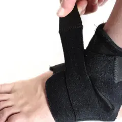 Dłoń zapina czarny stabilizator na kostce. Zapewnia wsparcie i ochronę podczas aktywności.