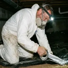Man i skyddsdräkt tätar krypgrund med plast och silvertejp för att motverka fukt.