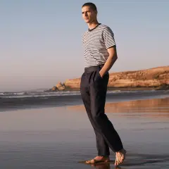 Mężczyzna w granatowych chinosach i pasiastym T-shircie spaceruje po plaży. Chinosy co to? To uniwersalne spodnie na każdą okazję.
