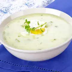 Kremowa zupa z sałaty z jajkiem i ziołami, podana w białej misce na niebieskim obrusie.