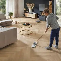 Frau wischt den hellen Designboden im Wohnzimmer. Ein Saugroboter ist im Hintergrund zu sehen.