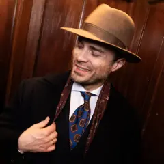 Elegancki mężczyzna w brązowym kapeluszu fedora, z krawatem i apaszką, poprawia marynarkę.