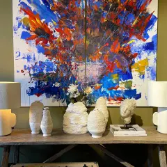 Dos lámparas sobremesa modernas blancas flanquean un cuadro abstracto vibrante. Jarrones texturizados y un busto completan la escena.