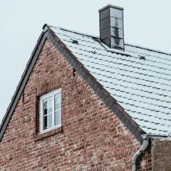 Ett tegeltak med snö och en mörk rökkanal skorsten mot en grå himmel.