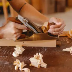 Main gauche tenant un rabot en laiton, créant des copeaux de bois. Une technique pour comment raboter du bois sans rabot.