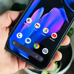 Dłoń trzyma smartfon z kolorową tapetą i ikonami aplikacji, w tym Sklep Play, Gmail, Zdjęcia i YouTube. Wygląda jak nowy **android 15**.