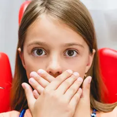 Przerażona dziewczynka zakrywa usta dłońmi, gdy dentysta przygotowuje się do podania znieczulenia. Wokół niej widać czerwony fotel dentystyczny i narzędzia.