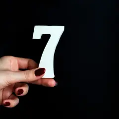 Dłoń z ciemnoczerwonym lakierem trzyma białą cyfrę 7, symbolizującą numerologiczną 7.