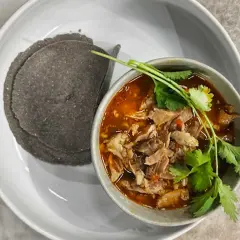 En skål med mustig el birria, toppad med koriander, serveras med mörka majstortillas på en vit tallrik.