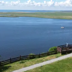 Widok na jezioro z pomostem i postacią, w oddali żaglówka. Krajobraz przypomina dawne grodzisko Lubin.