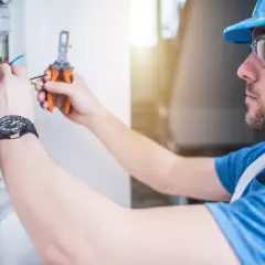 Elektryk przemysłowy w kasku i okularach ochronnych pracuje przy skrzynce z przewodami, używając obcinaczek.