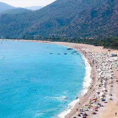 Tłum ludzi odpoczywa na piaszczystej plaży, kąpiąc się w turkusowym morzu. Widok na riwierę egejską z górzystym, zalesionym tłem.