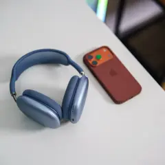 Niebieskie słuchawki AirPods Max i brązowy iPhone na białym biurku. Dowiedz się, jak połączyć AirPods z PC.