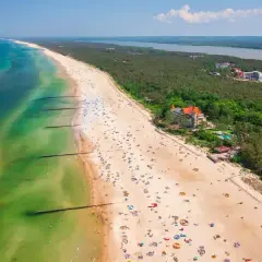 Słoneczna plaża nad Jeziorem Łebsko, pełna wypoczywających ludzi i zielony las. Atrakcje turystyczne w zasięgu wzroku.