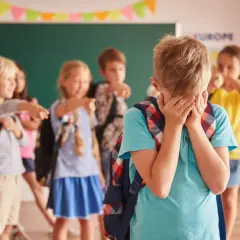 Chłopiec płacze, zakrywając twarz dłońmi, podczas gdy inne dzieci wskazują na niego palcami.
