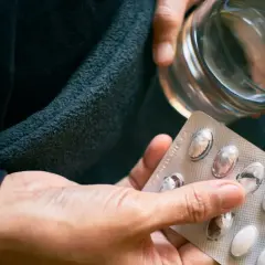 Dłoń trzyma blister z tabletkami i szklankę wody. Czy to próba zwalczenia nadmiaru witaminy B12?