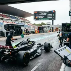 Coche de F1 en boxes durante entrenamientos. Un miembro del equipo guía al piloto mientras otros revisan tiempos en pantalla.