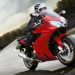 Motociclista en moto roja deportiva, con casco y equipo de cuero, inclinándose en una curva. El carenado de protección de la moto es aerodinámico y brillante.
