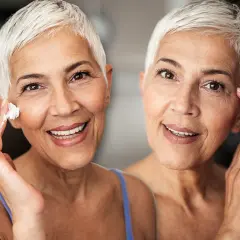 Zwei Frauen mit kurzen weißen Haaren lächeln. Eine trägt Creme auf ihre Wange auf, um trockene Haut im Alter zu bekämpfen.
