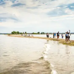Ludzie spacerują po piaszczystej mierzei Rewskiej, ciesząc się słońcem i wodą. Na horyzoncie widać łodzie i zielone drzewa.