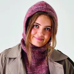 Ung kvinna med blå ögon bär en stickad balaclava i rosa och lila nyanser, med en beige trenchcoat.