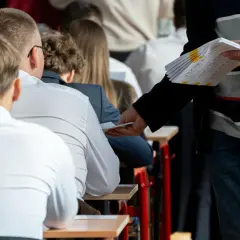 Nauczyciel rozdaje arkusze egzaminacyjne uczniom siedzącym w ławkach.