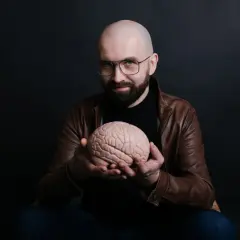 Łysy mężczyzna w okularach i brązowej kurtce trzyma w dłoniach model mózgu.