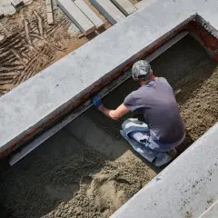 Budowlaniec wyrównuje masę, która będzie stanowiła jaki beton na fundament. Widać ceglane ściany i betonowe elementy konstrukcji.