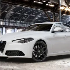 Biała Alfa Romeo Giulia Veloce z czarnymi felgami stoi w opuszczonej hali przemysłowej, gdzie światło przebija się przez dach.