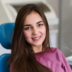 Uśmiechnięta nastolatka z kolorowym aparatem ruchomym na zębach siedzi na niebieskim fotelu dentystycznym.